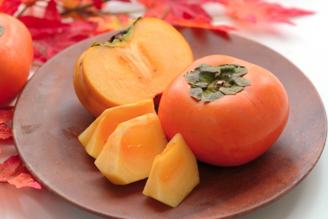 Japanese persimmons, including a whole fruit and sliced pieces, displayed on a wooden plate