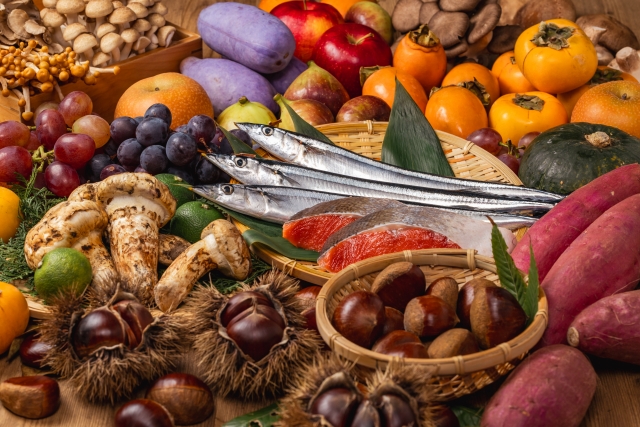 A variety of Japanese autumn foods, including Matsutake mushrooms, Sanma fish, chestnuts, sweet potatoes, and seasonal fruits, arranged on a table