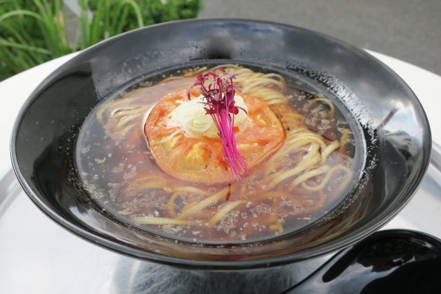 A bowl of vegan ramen with a clear broth, topped with a slice of grilled tomato, fresh greens, and purple sprouts