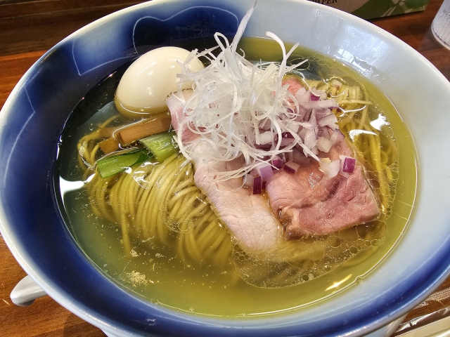 A bowl of shio ramen with a clear, light broth, topped with tender slices of pork, a soft-boiled egg, and finely chopped green onions